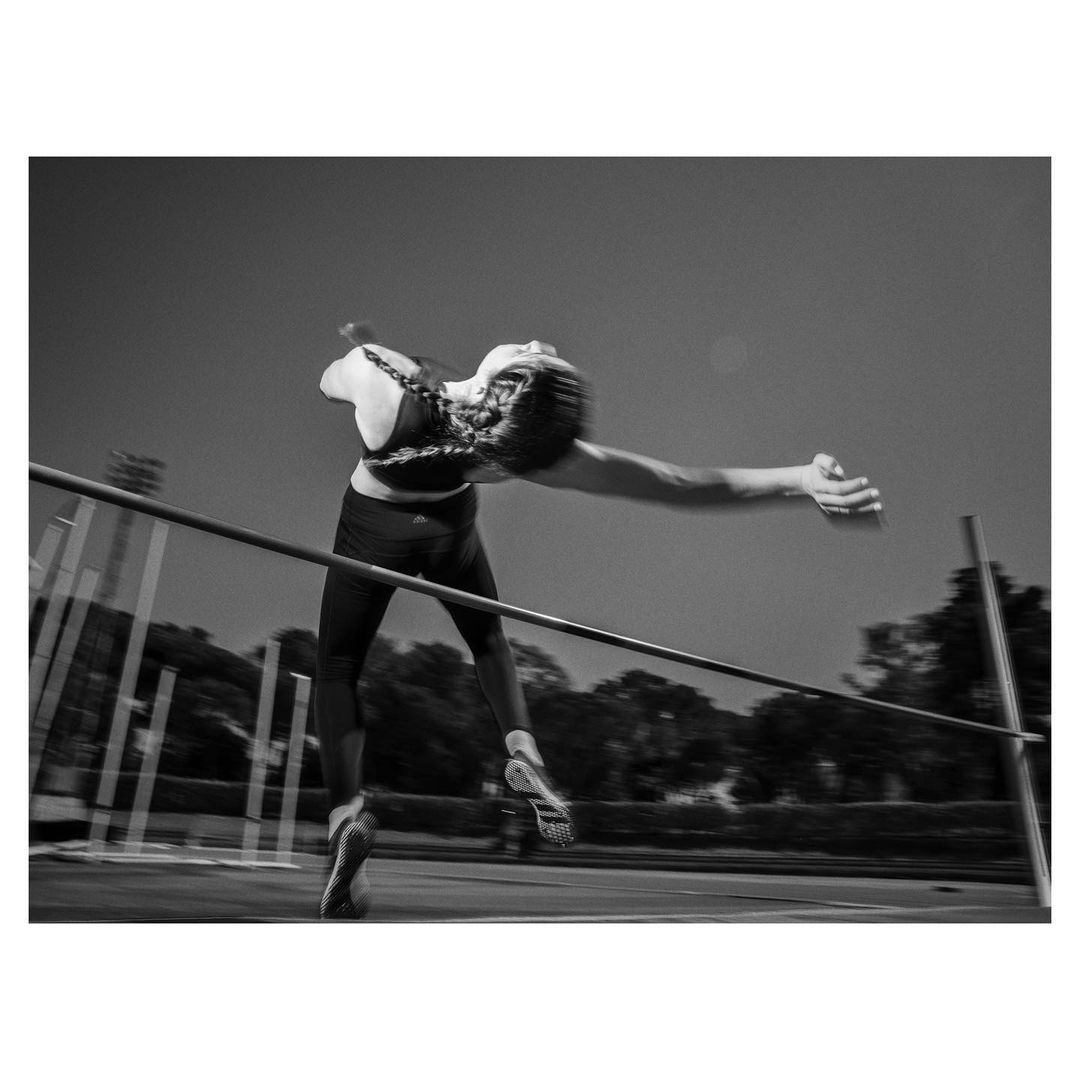 Monochrome photo of a female high jumper in mid-air, clearing the bar at an outdoor track and field venue on a sunny day.