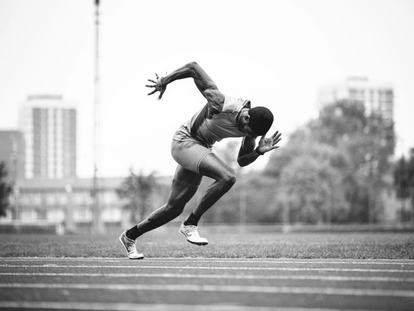 Monochrome image of a muscular runner sprinting on a track outdoors, showcasing intense motion and athletic power.