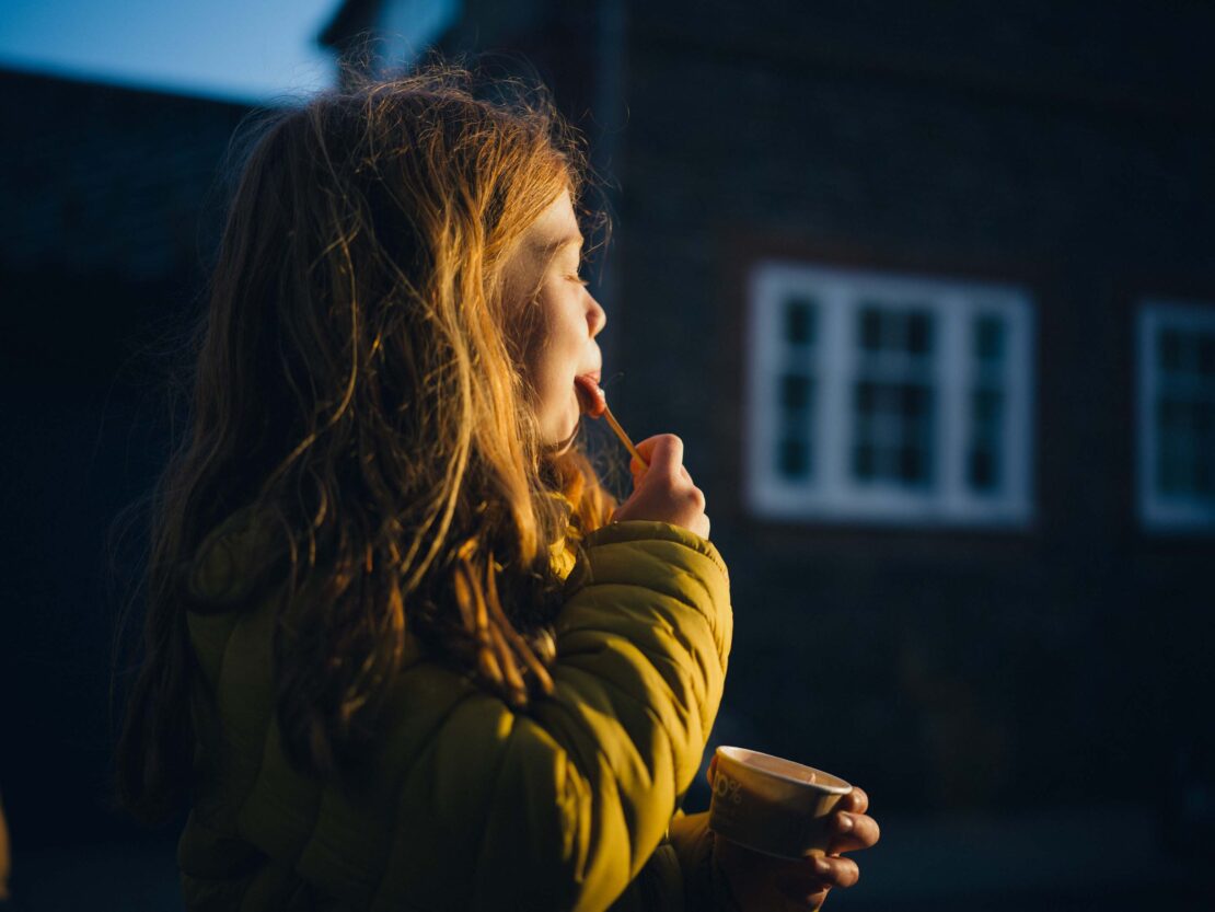 Young girl with red hair enjoys a sweet treat outdoors. She's wearing a yellow puffer jacket, catching the sunlight in the evening.