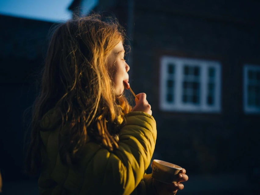 Young girl with red hair enjoys a sweet treat outdoors. She's wearing a yellow puffer jacket, catching the sunlight in the evening.