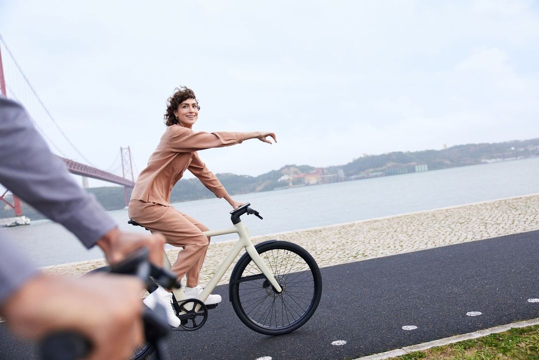Woman in a tan suit rides a bicycle on a path near a bridge and river, smiling as she gestures, capturing an active outdoor scene.