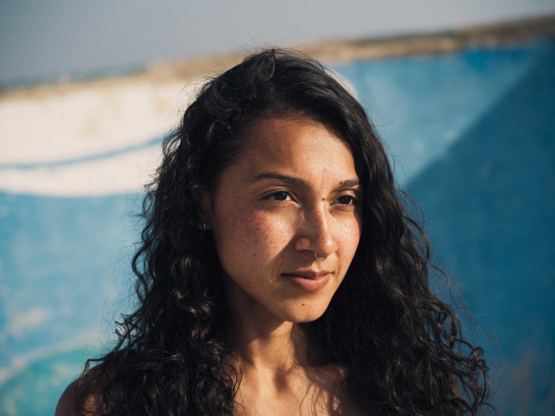 Close-up portrait of a woman with dark curly hair and a nose ring outdoors against a blue and white background by Paul Calver.