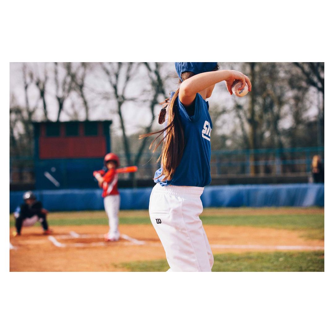 Young girl pitches baseball on sunny outdoor field with batter and catcher in background, wearing team uniform, white pants, and blue cap.