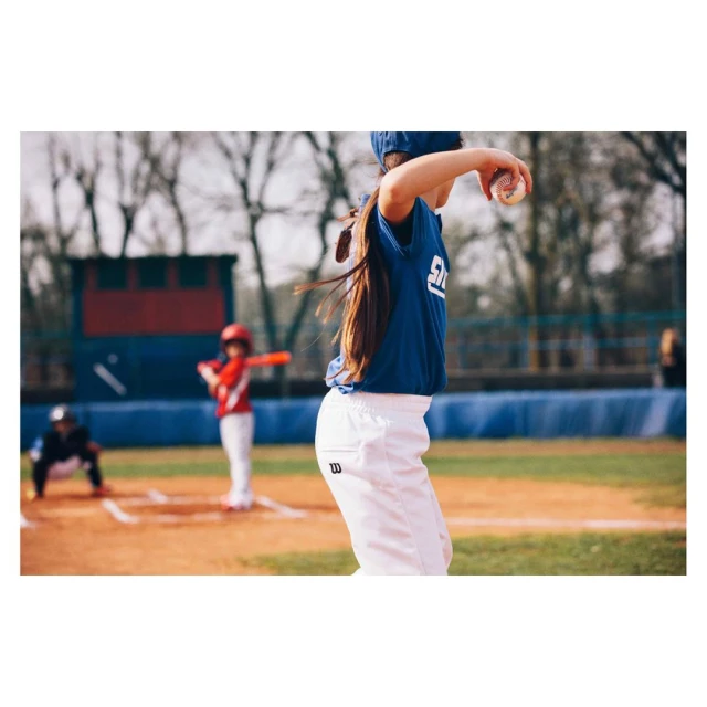 Young girl pitches baseball on sunny outdoor field with batter and catcher in background, wearing team uniform, white pants, and blue cap.