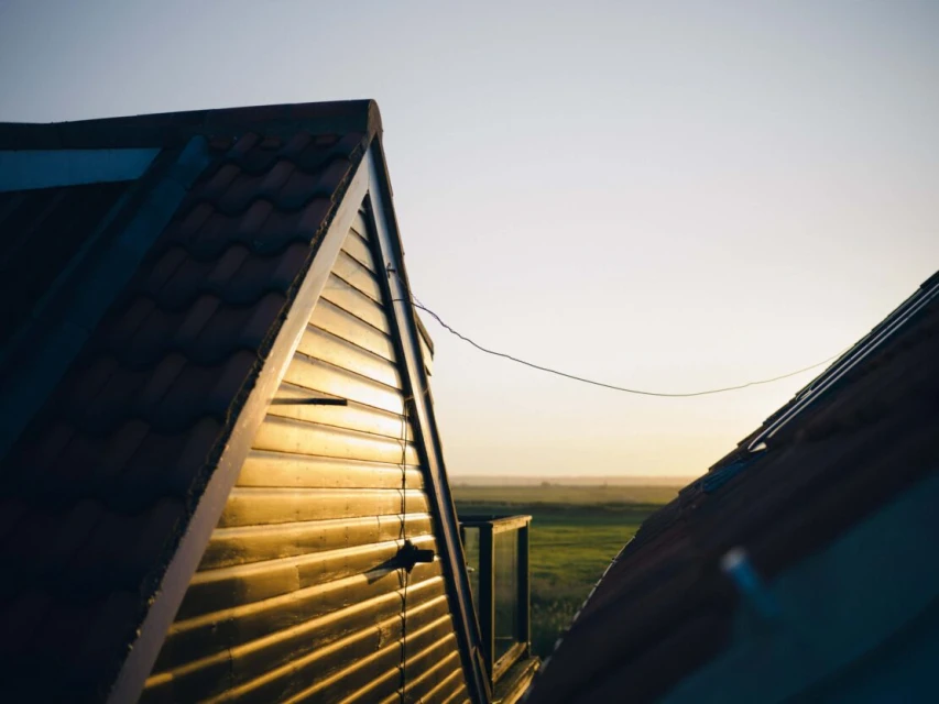 View of rooftop and green field under a bright sky at sunset from a low angle, with warm sunlight reflecting off the roof siding.