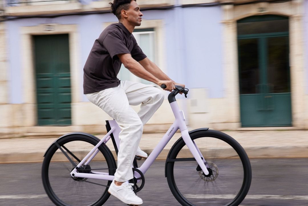 A man rides a light purple Cowboy brand bicycle on a city street on a sunny day. He wears a t-shirt, white pants, and white sneakers.