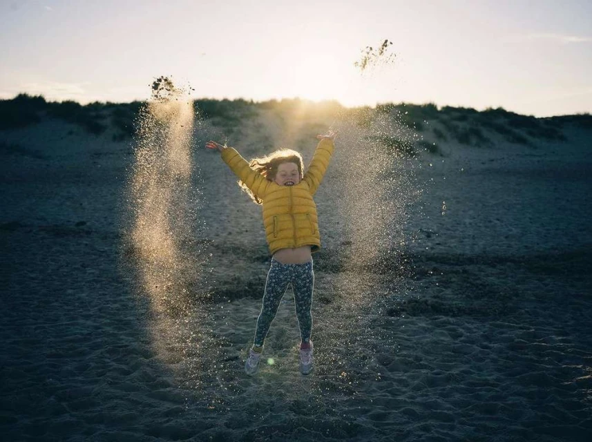 A child in a yellow jacket jumps in the air on a sandy beach, throwing sand with outstretched arms towards a bright, sunny sky.