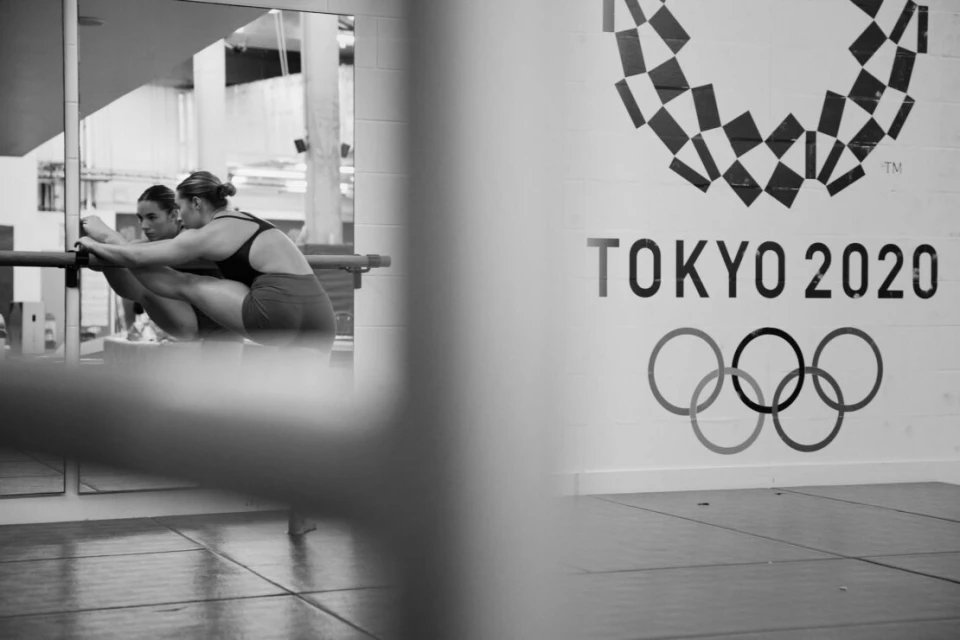 Female athlete stretching at ballet barre in monochrome studio with Tokyo 2020 Olympics logo on the wall in the background.