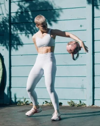 Athletic woman with short blonde hair wearing white activewear holding a kettlebell outdoors by a blue wall