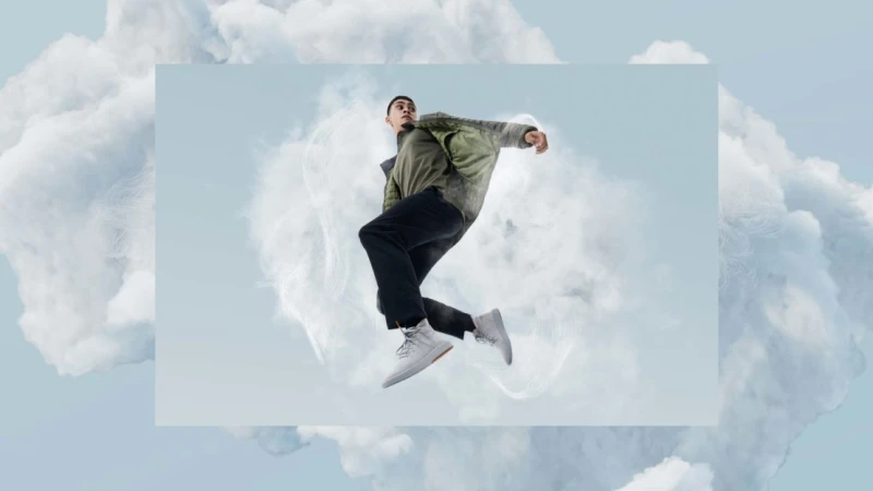 A man in a green jacket and dark trousers leaps mid-air inside a framed studio shot surrounded by large white clouds.