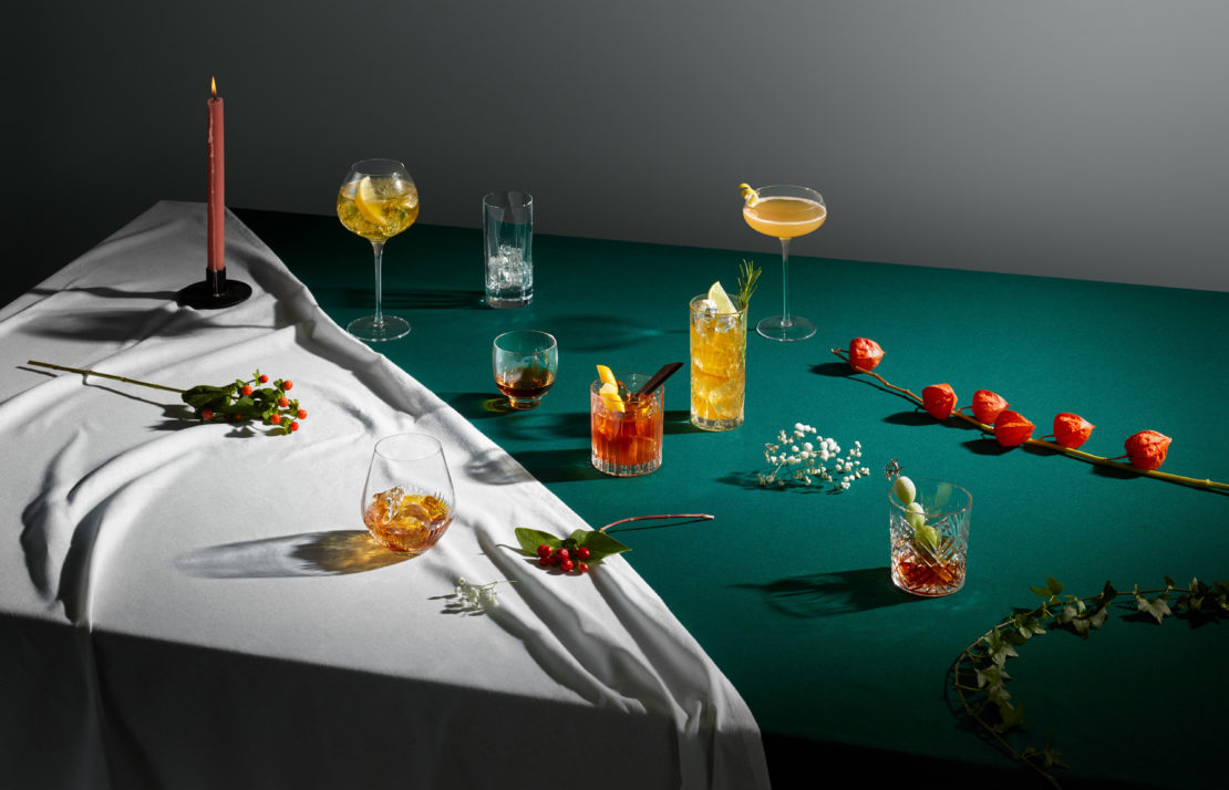 A table setup showcasing an assortment of colorful cocktails in glassware, complemented by botanical garnishes and moody, controlled lighting.