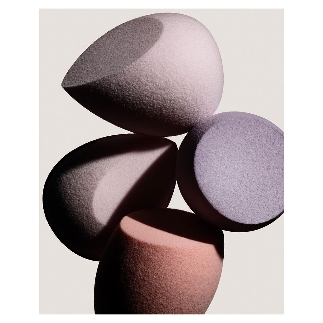 Close-up still life of four pastel-colored makeup blending sponges in shades of lavender, pink, and beige, on a neutral surface.