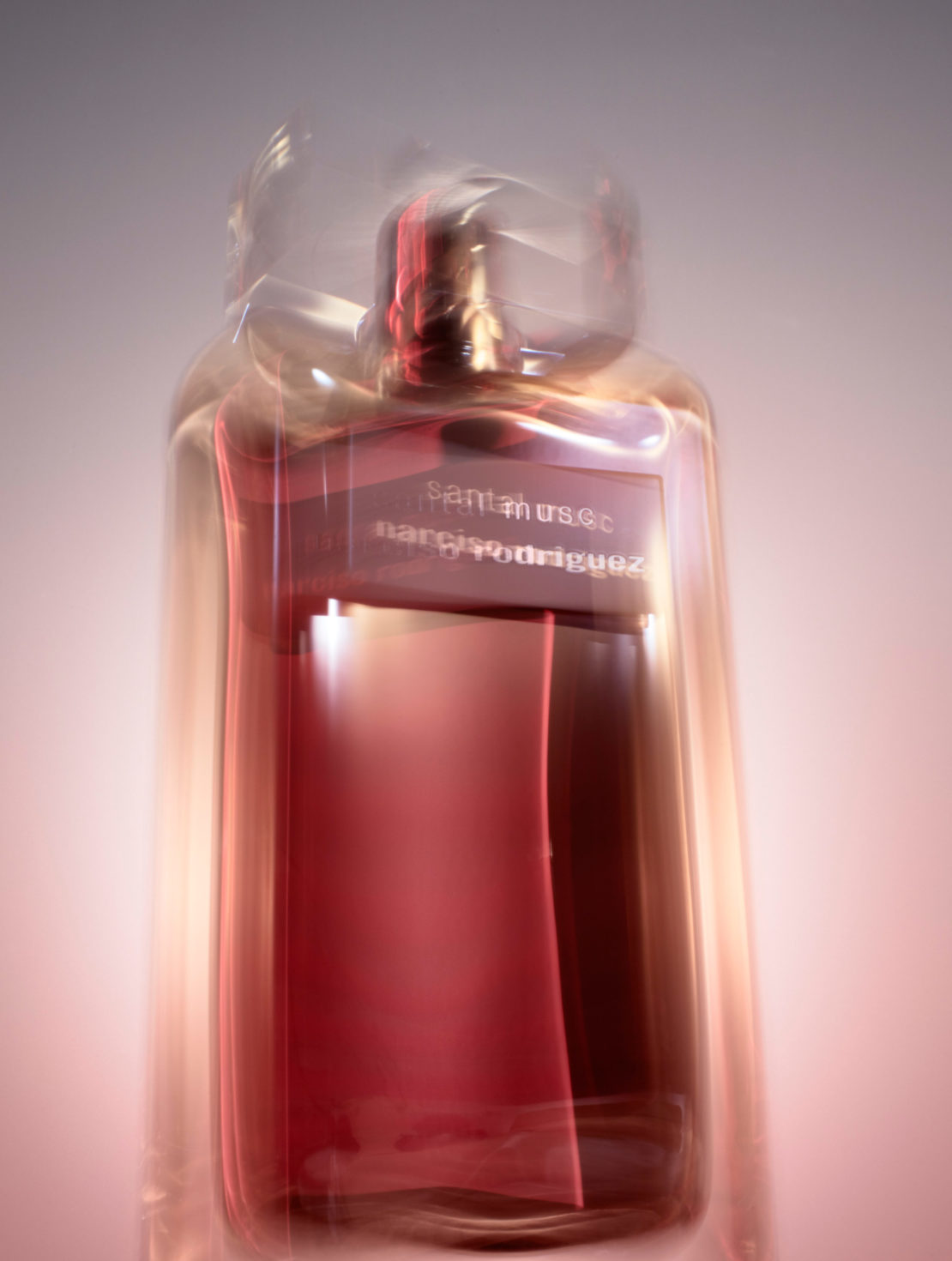 Close-up studio shot of a blurred perfume bottle with a red label. The bottle is clear with a red tint and soft lighting.