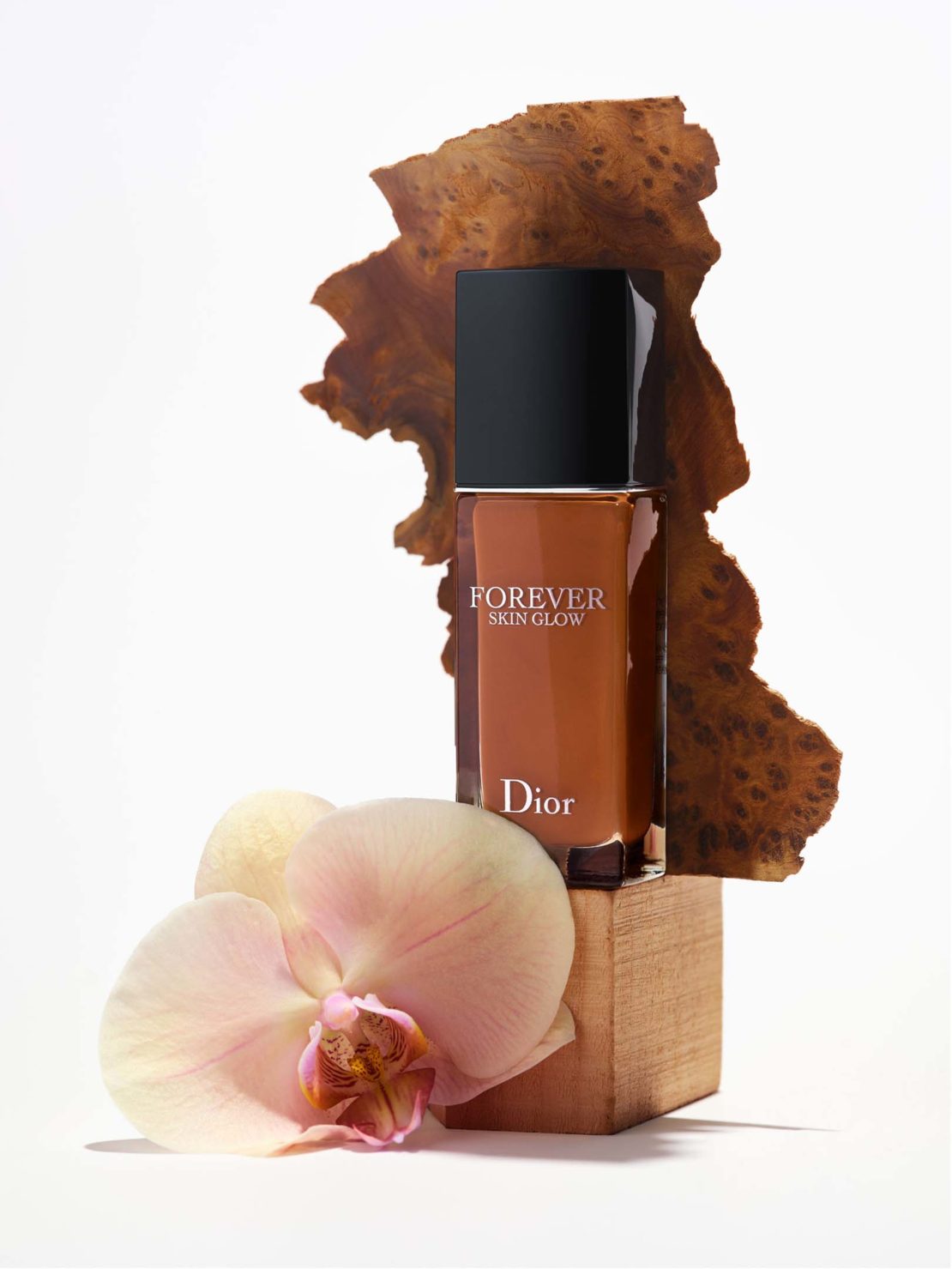 Dior Forever Skin Glow foundation bottle on a wooden block, next to an orchid, set against a clean white background.