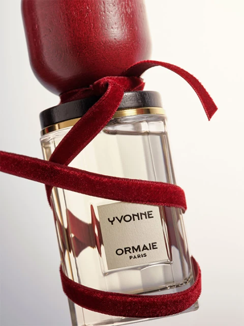 Close-up of a glass perfume bottle with a dark wood cap and a red velvet ribbon against a plain white backdrop in a studio setting.