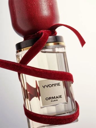 Close-up of a glass perfume bottle with a dark wood cap and a red velvet ribbon against a plain white backdrop in a studio setting.