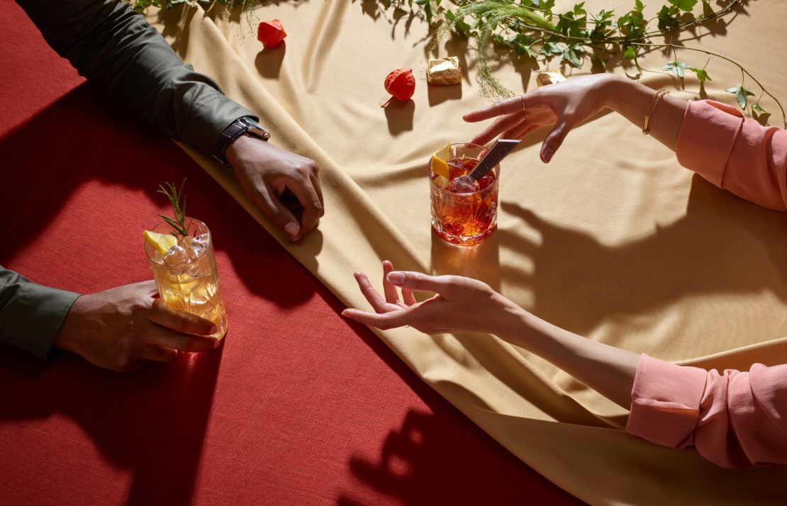 Two cocktails served on a draped table with hands reaching for them. One light amber and the other dark amber.
