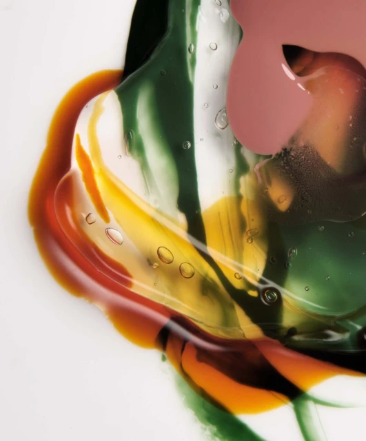 Close-up of liquid cosmetic swatches in shades of amber, green, yellow, and rose on a white surface, creating an abstract pattern.