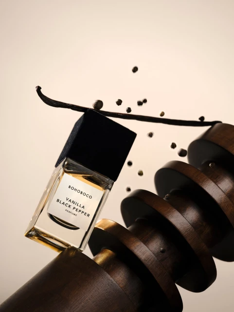BOHOBOCO Vanilla Black Pepper perfume bottle with vanilla bean and black peppercorns, balanced on wood circles, studio shot.