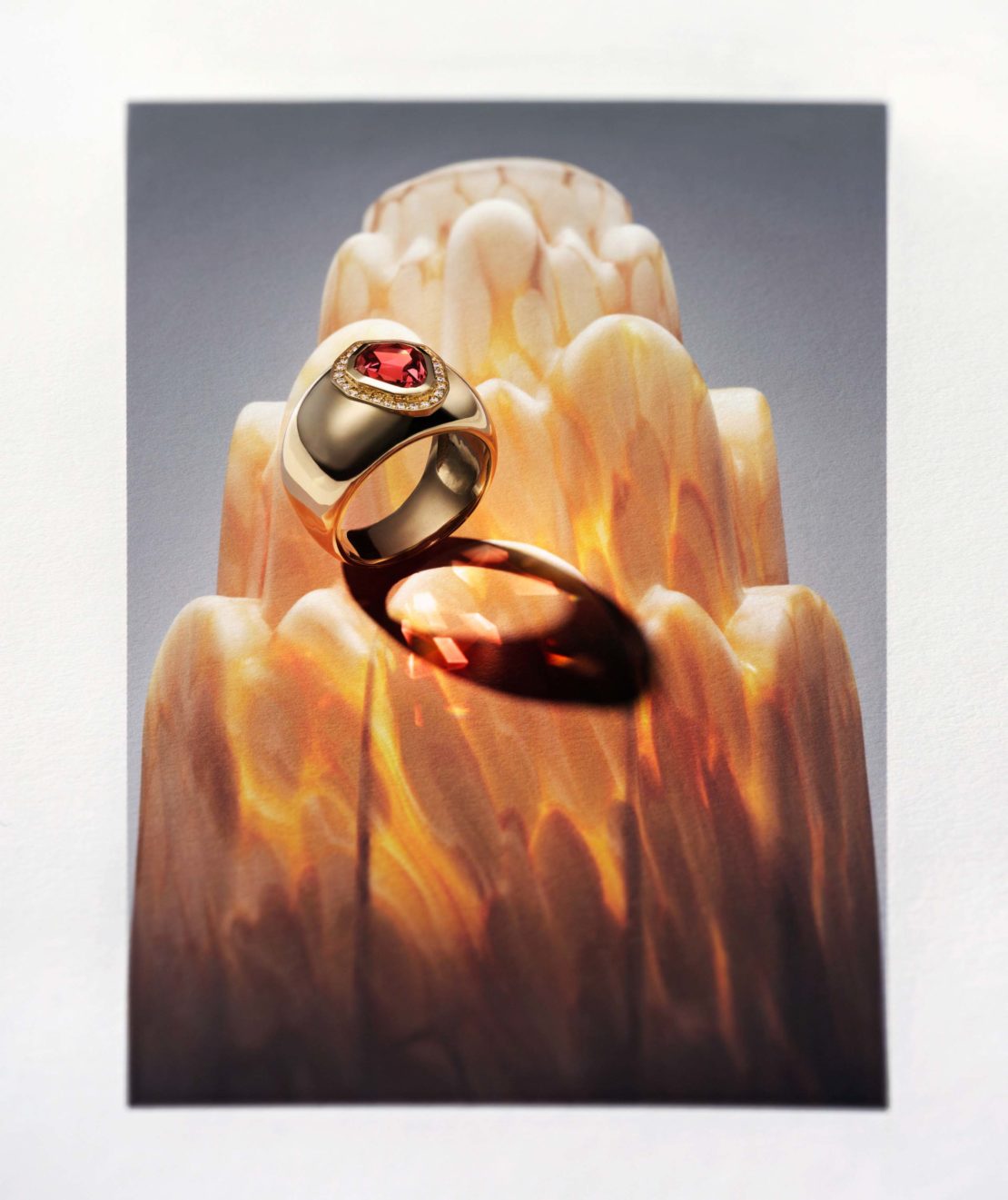 Gold ring featuring a red gemstone, displayed on a decorative stand with layered patterns, illuminated with soft lighting, studio shot.