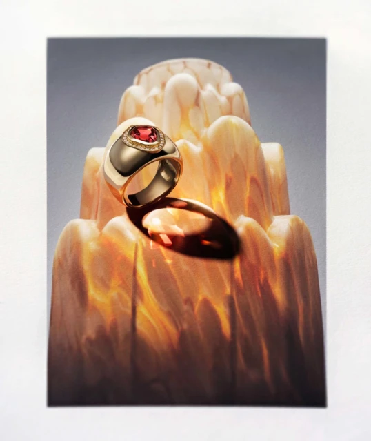 Gold ring featuring a red gemstone, displayed on a decorative stand with layered patterns, illuminated with soft lighting, studio shot.