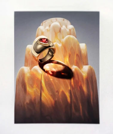 Gold ring featuring a red gemstone, displayed on a decorative stand with layered patterns, illuminated with soft lighting, studio shot.
