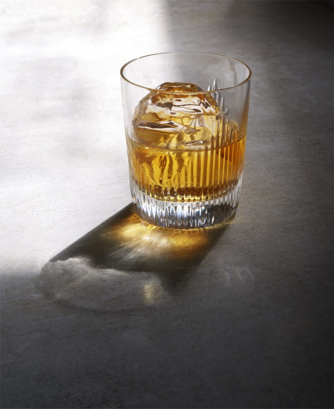 Crystal whiskey glass with large round ice cube and amber liquid on a grey stone surface with strong directional light shadow