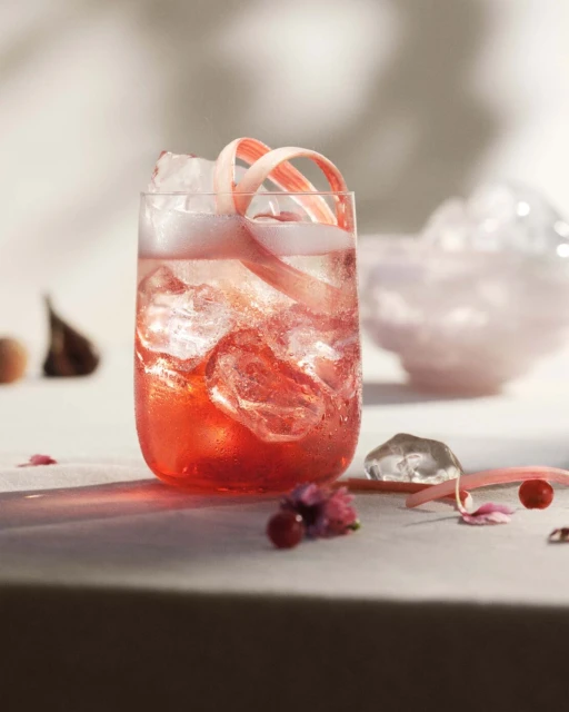 A glass filled with a pink drink, ice, and a decorative rhubarb curl, set against a bright background with a decorative glass.