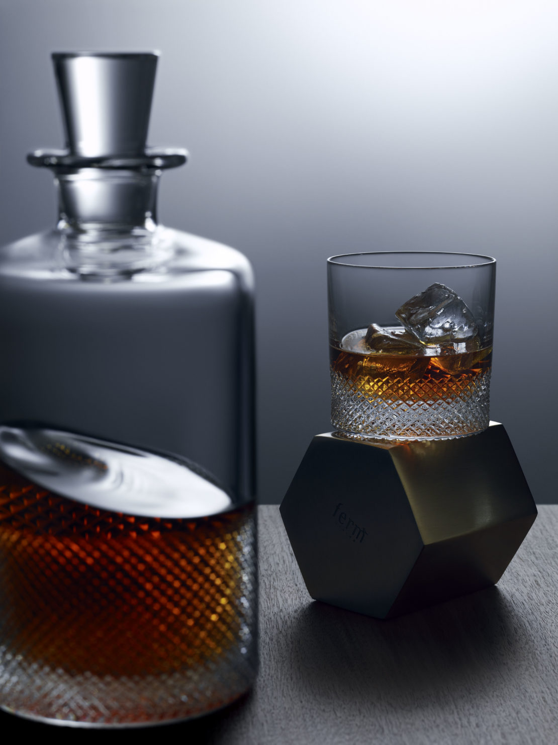 Crystal whiskey decanter and glass with ice on dark wood surface, illuminated by dramatic lighting against a neutral background.