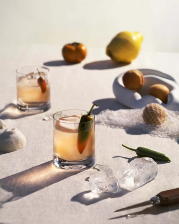 Close-up of two cocktails with ice and pepper garnish, next to fruits, white decorative objects, and crushed ice on a white surface.