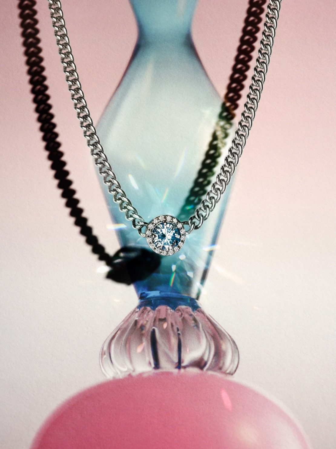 Elegant silver chain necklace with a blue gemstone pendant, displayed against a soft pink backdrop with blue glass vase elements.