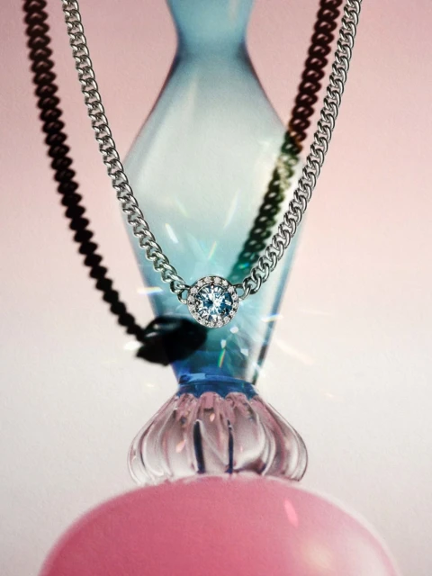 Elegant silver chain necklace with a blue gemstone pendant, displayed against a soft pink backdrop with blue glass vase elements.