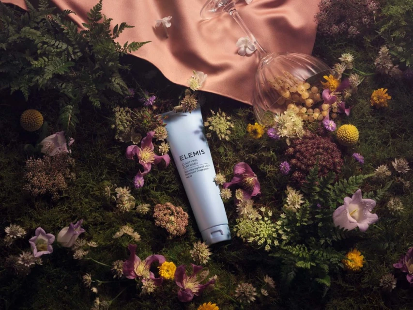 Elemis Clarifying Clay Cleanser lies on moss amongst flowers and foliage. A glass and peach-toned cloth are in the background.