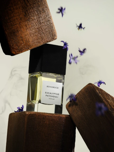 Perfume bottle with black cap on wood blocks, surrounded by purple flowers, minimalist studio product shot, beige background.