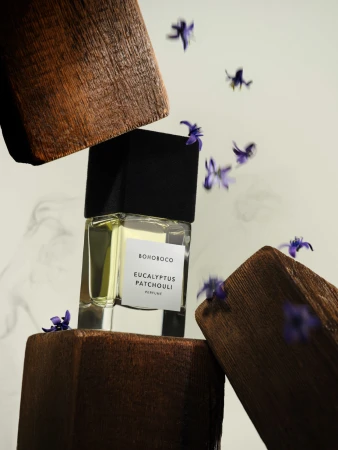 Perfume bottle with black cap on wood blocks, surrounded by purple flowers, minimalist studio product shot, beige background.