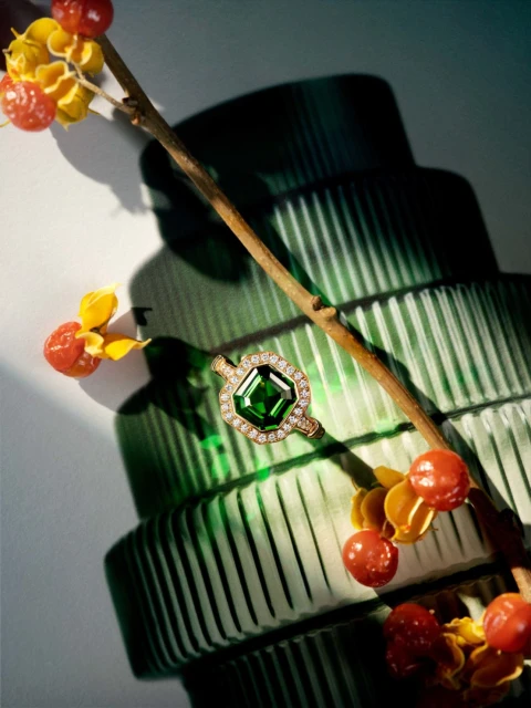 Emerald-cut green gemstone ring surrounded by small diamonds on a branch with red berries, next to a green glass vase.