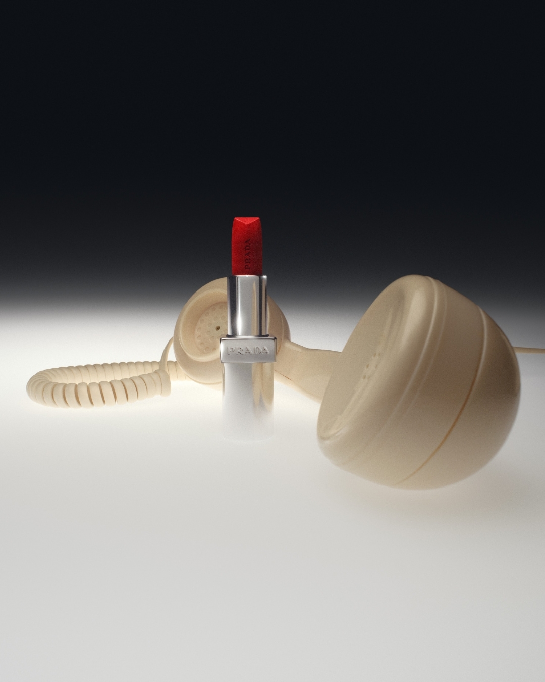Product shot featuring red lipstick with the brand Prada on its silver case, next to a vintage beige landline telephone on a light surface.