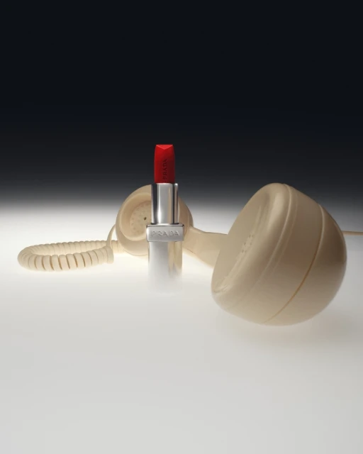 Product shot featuring red lipstick with the brand Prada on its silver case, next to a vintage beige landline telephone on a light surface.