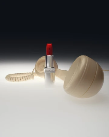 Product shot featuring red lipstick with the brand Prada on its silver case, next to a vintage beige landline telephone on a light surface.