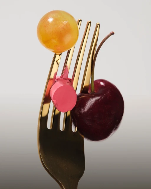 Close-up of a golden fork holding a green grape, a pink lipstick bullet, and a red cherry, all against a neutral background.