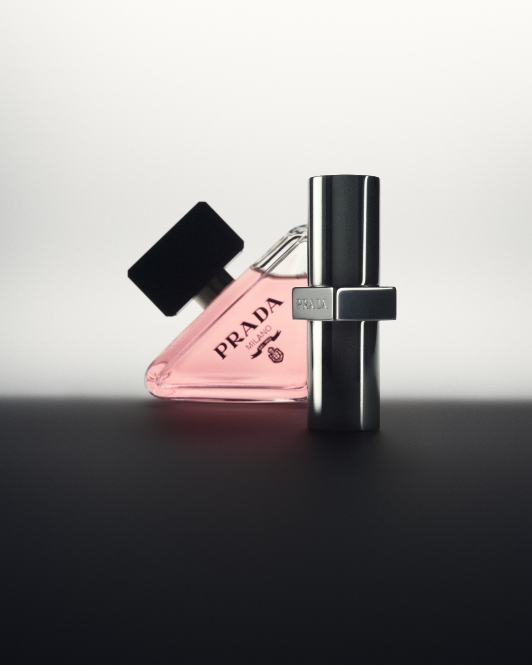 Product shot of Prada Paradoxe fragrance, featuring a triangle-shaped bottle with pink liquid and a silver metal bottle, against a graduated background.