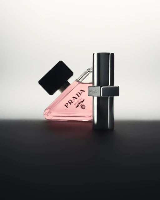 Product shot of Prada Paradoxe fragrance, featuring a triangle-shaped bottle with pink liquid and a silver metal bottle, against a graduated background.