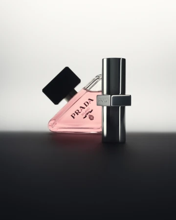 Product shot of Prada Paradoxe fragrance, featuring a triangle-shaped bottle with pink liquid and a silver metal bottle, against a graduated background.