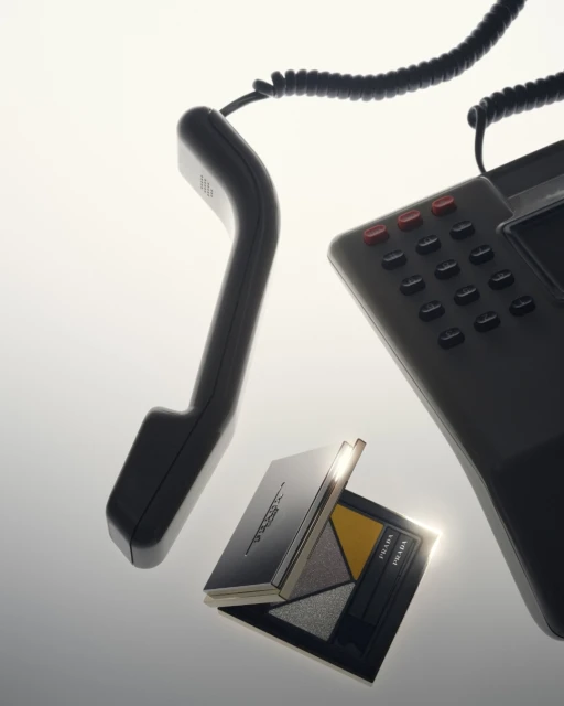 Dark grey cordless phone receiver and phone base next to open Prada eyeshadow palette, against a bright gradated white background.