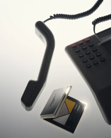 Dark grey cordless phone receiver and phone base next to open Prada eyeshadow palette, against a bright gradated white background.