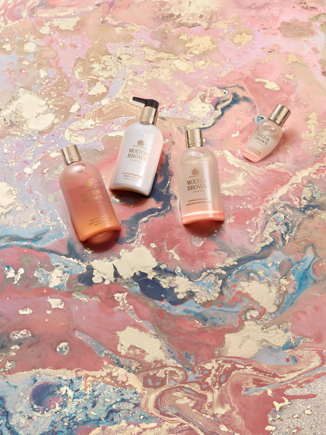 Four Molton Brown bath and body product bottles arranged on a pink, white, and blue marbled surface in a beauty product shot.