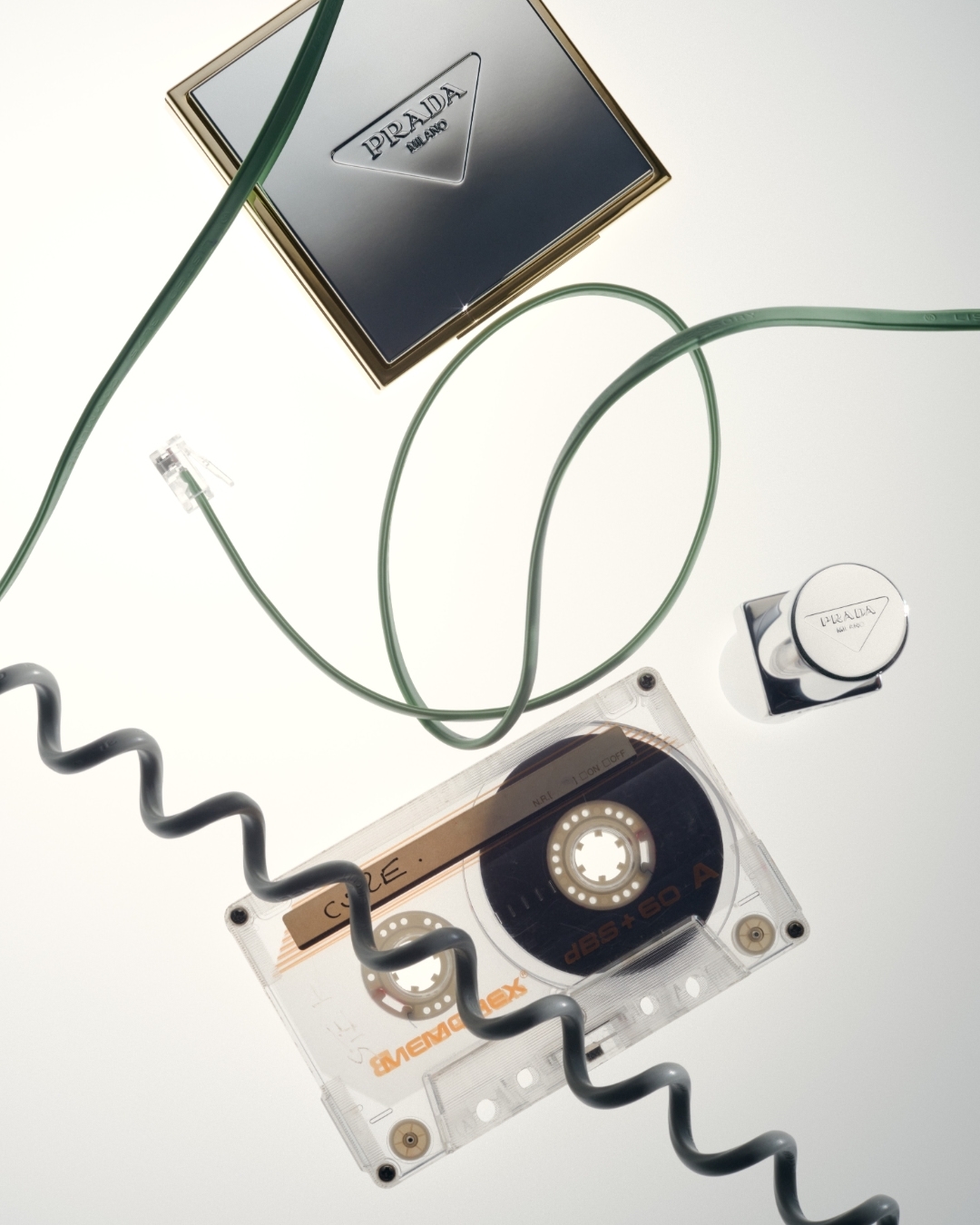 Overhead shot of Prada compacts, a cassette tape, and green and black cables on a white surface, emphasizing retro tech and luxury beauty.