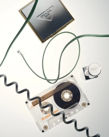 Overhead shot of Prada compacts, a cassette tape, and green and black cables on a white surface, emphasizing retro tech and luxury beauty.
