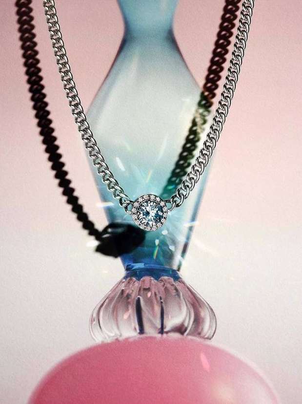 Close-up of a silver chain necklace with a diamond pendant draped over a blue glass vase, set against a pink backdrop.