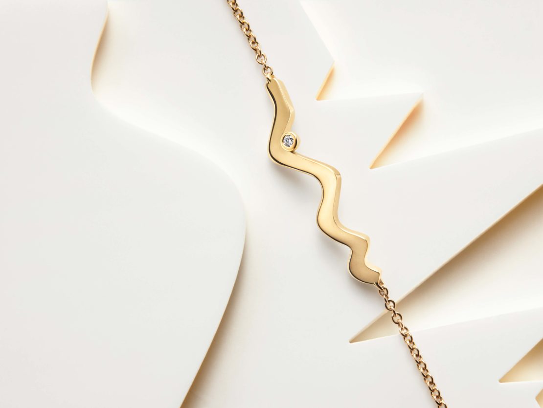 Gold necklace with a delicate chain and a wave-shaped pendant featuring a small diamond, set against a clean, bright paper surface.